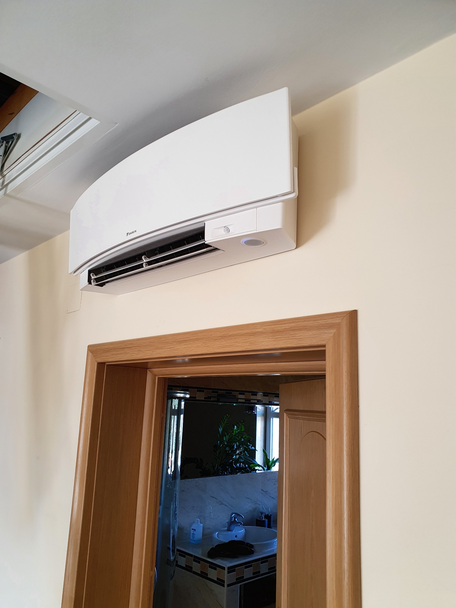 Biały klimatyzator marki Daikin zamontowany nad drewnianą futryną drzwi do łazienki z widocznym fragmentem umywalki i lustra.