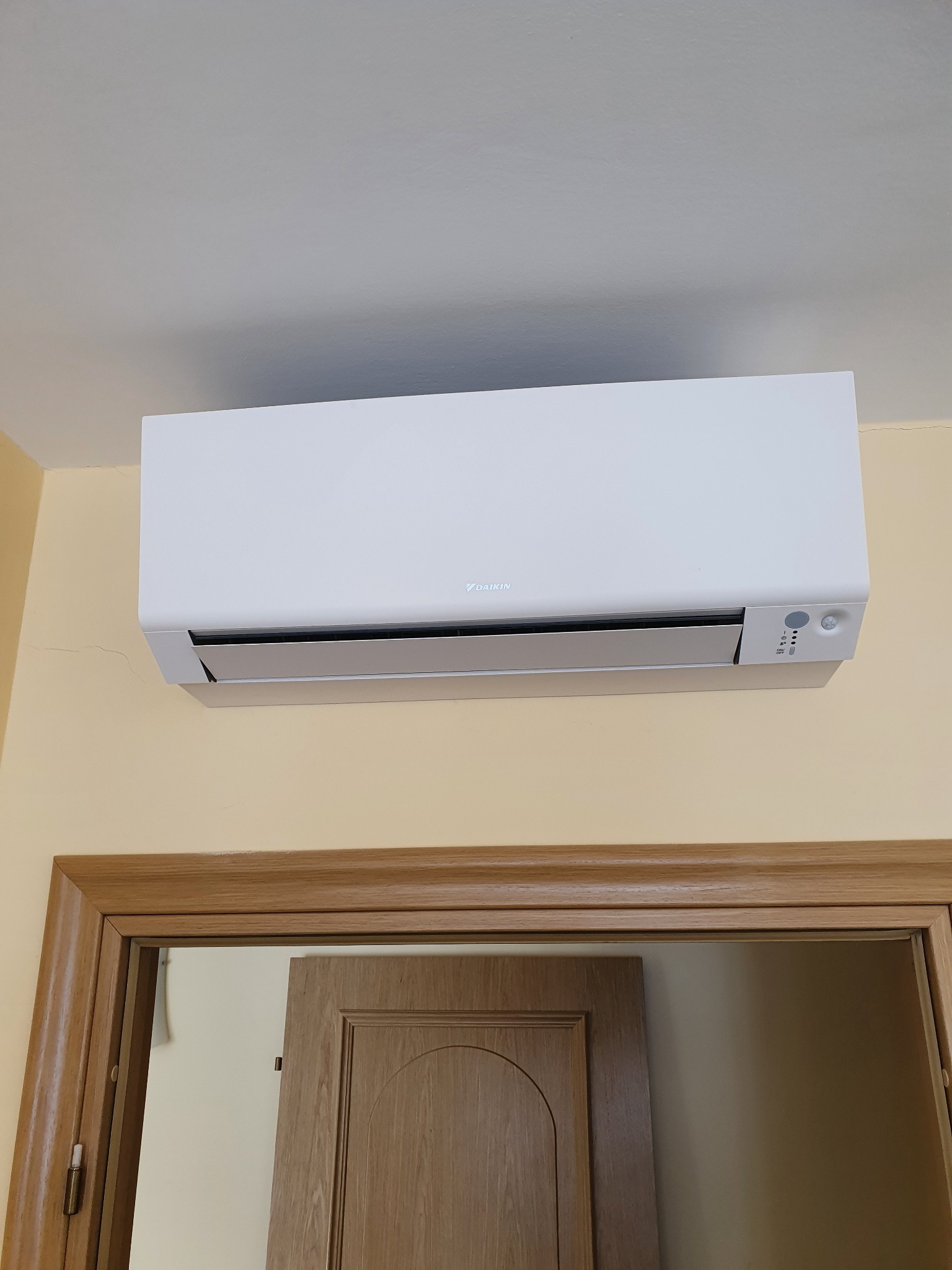 Biały klimatyzator Daikin zamontowany na ścianie nad drewnianą futryną drzwiową w pomieszczeniu o kremowych ścianach.