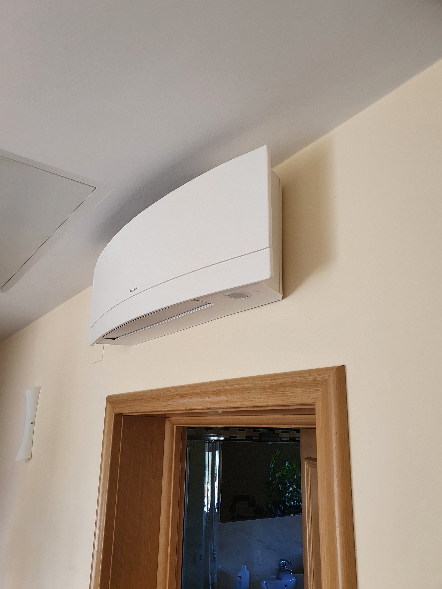 Biały klimatyzator Daikin zamontowany na ścianie nad drewnianą futryną drzwi do łazienki z widoczną umywalką i rośliną w tle.