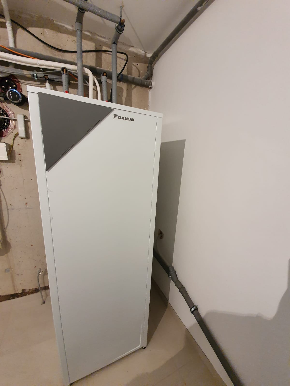 Biała pompa ciepła Daikin z szarym panelem na tle surowej ściany z rurami instalacji grzewczej i fragmentem białej ściany, widok z boku.