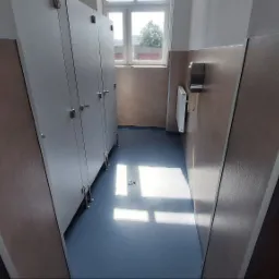 Wnętrze szatni z rzędem białych szafek, niebieską wykładziną podłogową i brązowymi panelami ściennymi, oświetlone naturalnym światłem wpadającym przez okno.