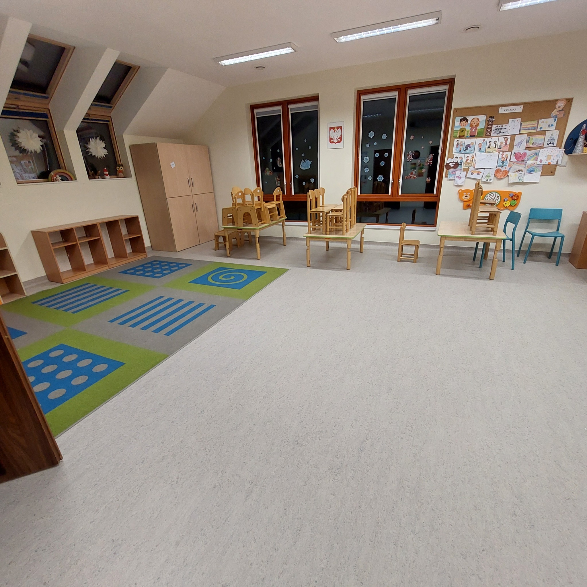 Sala przedszkolna z wykładziną dywanową w geometryczne wzory, drewnianymi meblami i dekoracjami okiennymi.