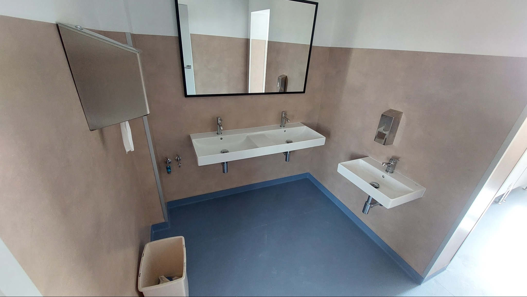 Łazienka publiczna z niebieską podłogą z wykładziny PCV, dwoma umywalkami i dużym lustrem w czarnej ramie.