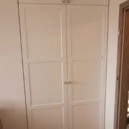 Biała szafa z czterema drzwiami, z panelami i okrągłymi gałkami, sięgająca od podłogi do sufitu, na drewnianej podłodze.