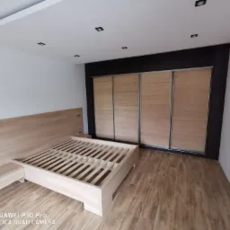 Sypialnia w trakcie wykańczania: rama łóżka z drewnianym stelażem, szafka nocna i przesuwne drzwi szafy wnękowej z jasnego drewna na tle białych ścian i drewnopodobnej podłogi.