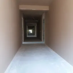 Długi korytarz w stanie surowym po nałożeniu gładzi, widok perspektywiczny z wyraźnym zbieganiem się linii w kierunku okna na końcu, widoczne wystające przewody elektryczne pod sufitem.