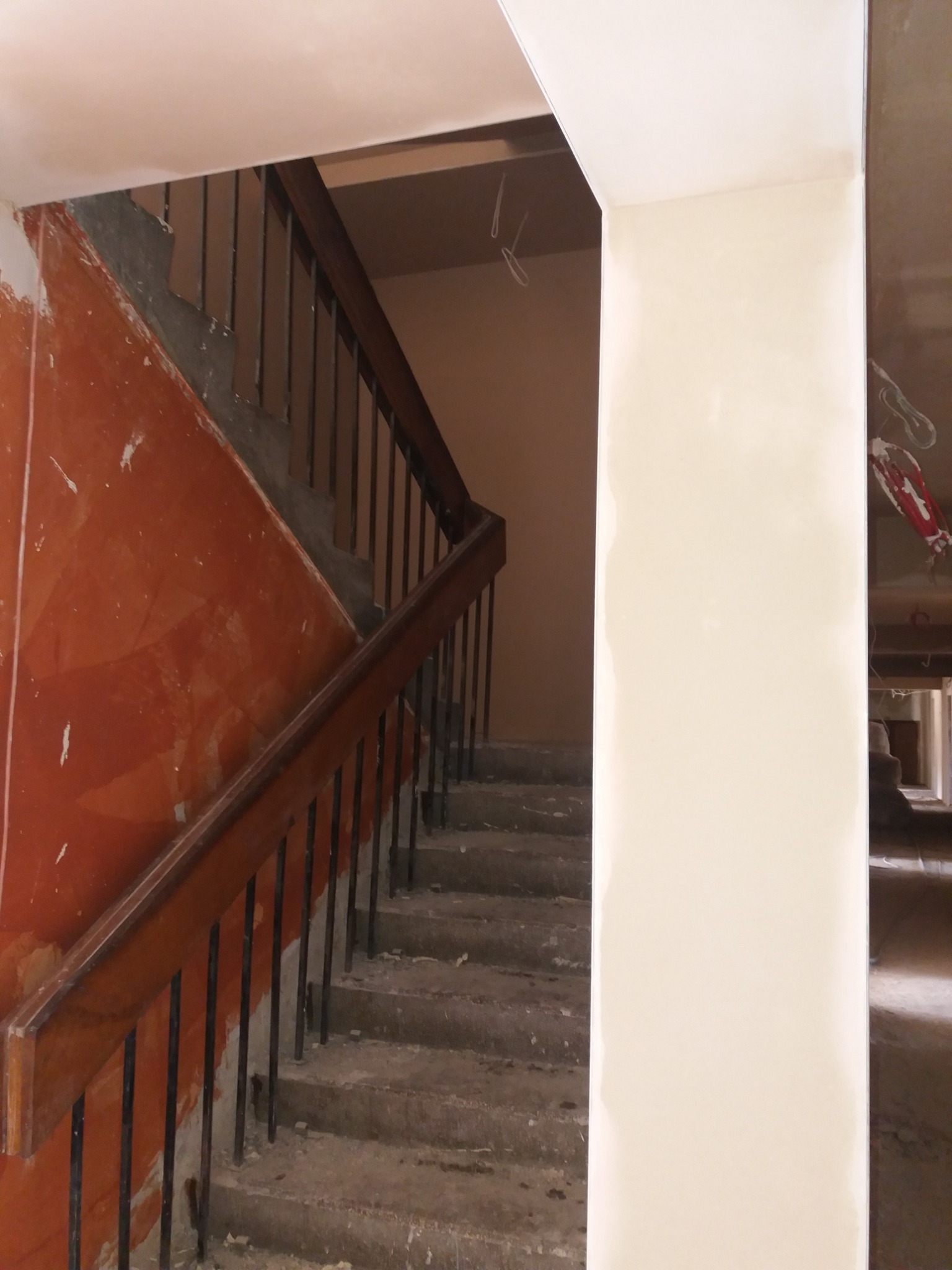Klatka schodowa w trakcie prac wykończeniowych: ściany częściowo pokryte nową, jasną powłoką, widoczne niedoskonałości i ślady po wcześniejszych pracach, surowe schody z metalową balustradą.