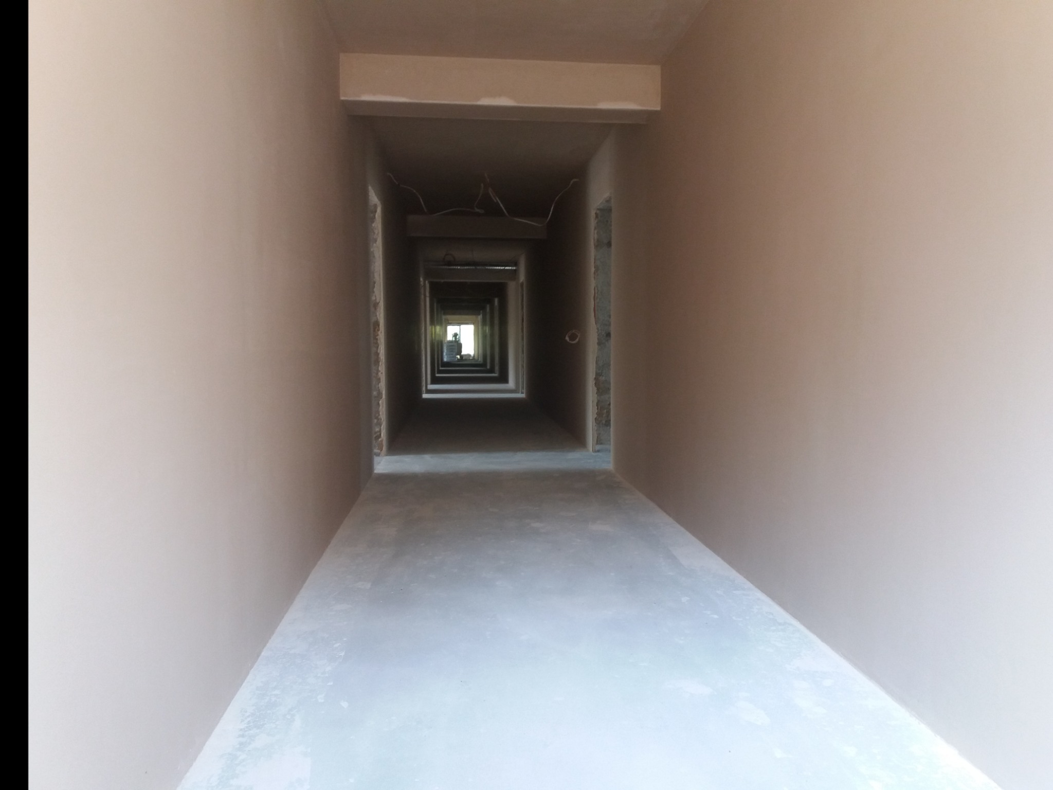 Długi korytarz w stanie surowym po nałożeniu gładzi, widok perspektywiczny z wyraźnym zbieganiem się linii w kierunku okna na końcu, widoczne wystające przewody elektryczne pod sufitem.