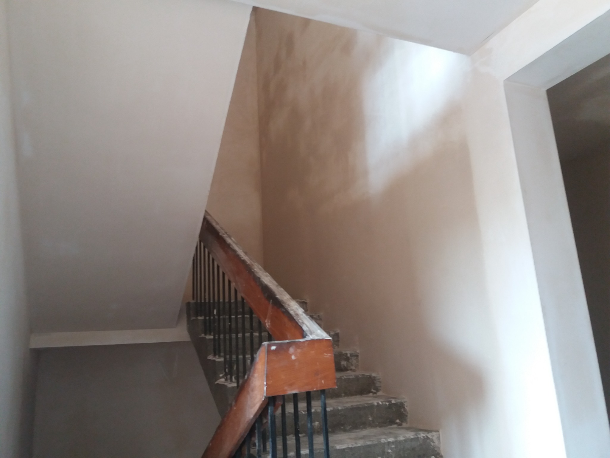 Świeżo otynkowana klatka schodowa z drewnianą balustradą, widoczne niedoskonałości tynku przed malowaniem, surowy beton schodów.