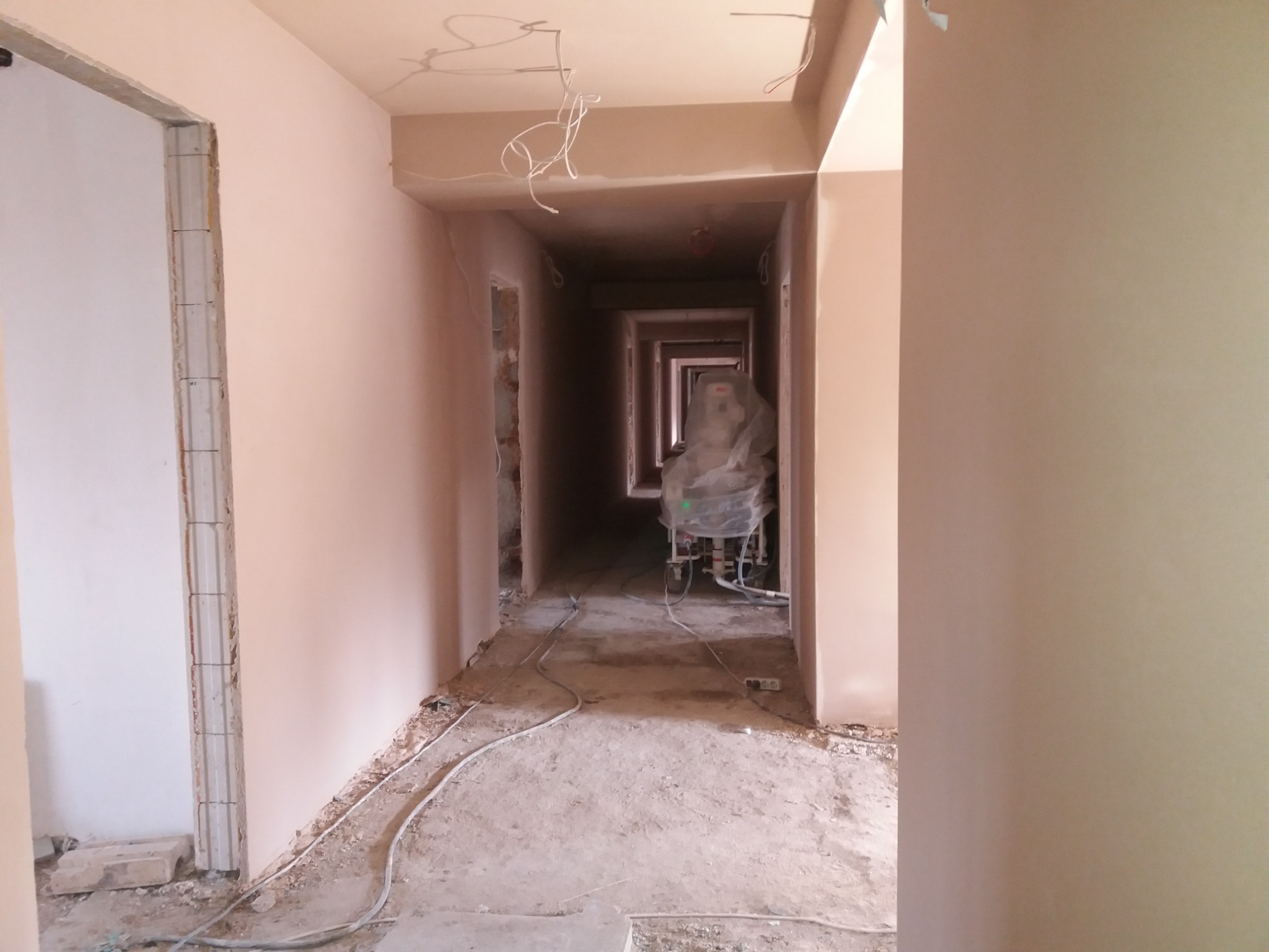 Korytarz w trakcie prac tynkarskich maszynowych; widoczna maszyna do tynkowania owinięta folią ochronną na końcu długiego korytarza oraz świeżo nałożone tynki na ścianach.