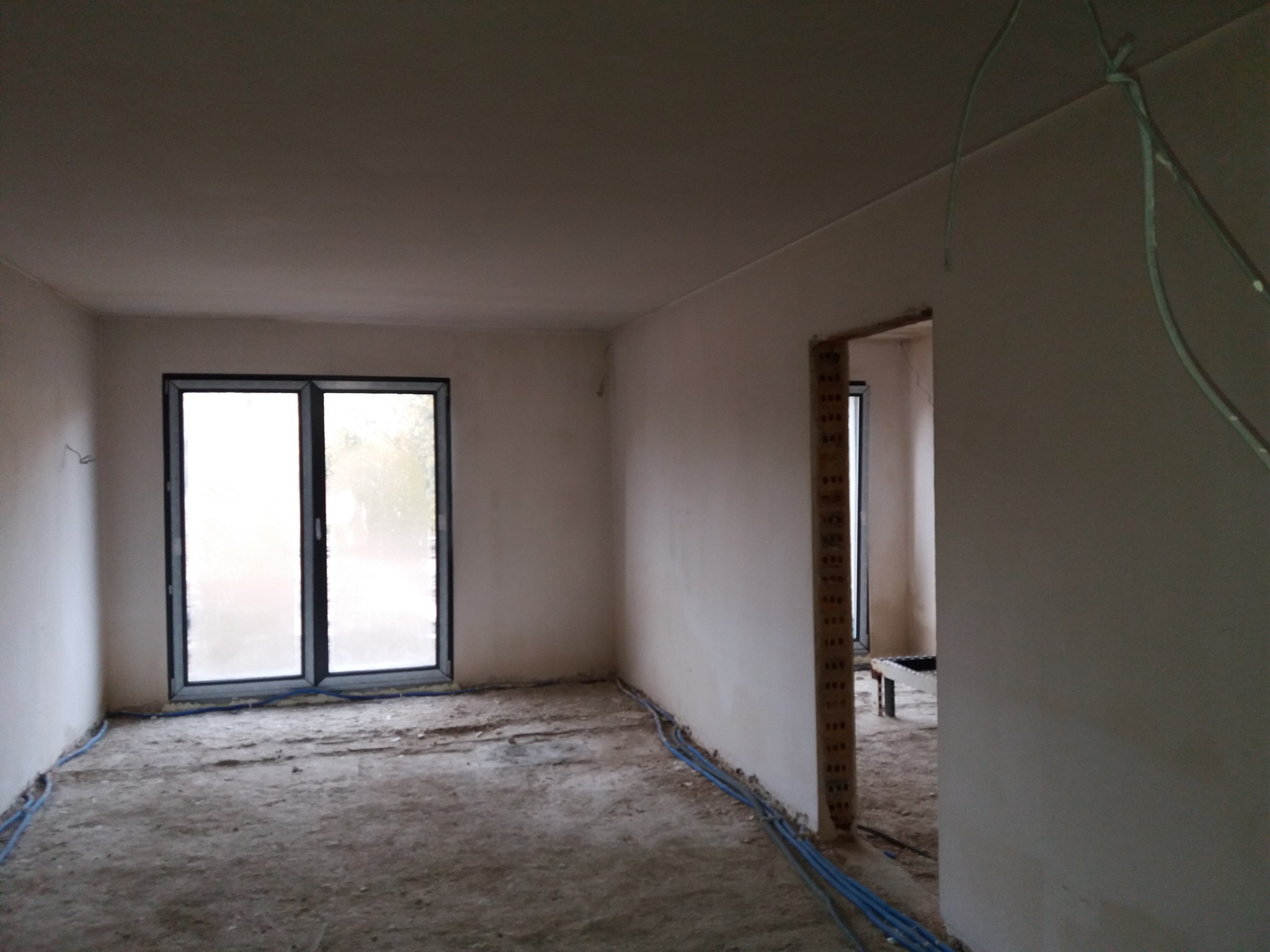 Wnętrze surowego pomieszczenia po nałożeniu tynku maszynowego, widoczne okno balkonowe z czarnymi ramami, niebieskie rurki instalacyjne na podłodze i fragment otworu drzwiowego z widoczną cegłą.