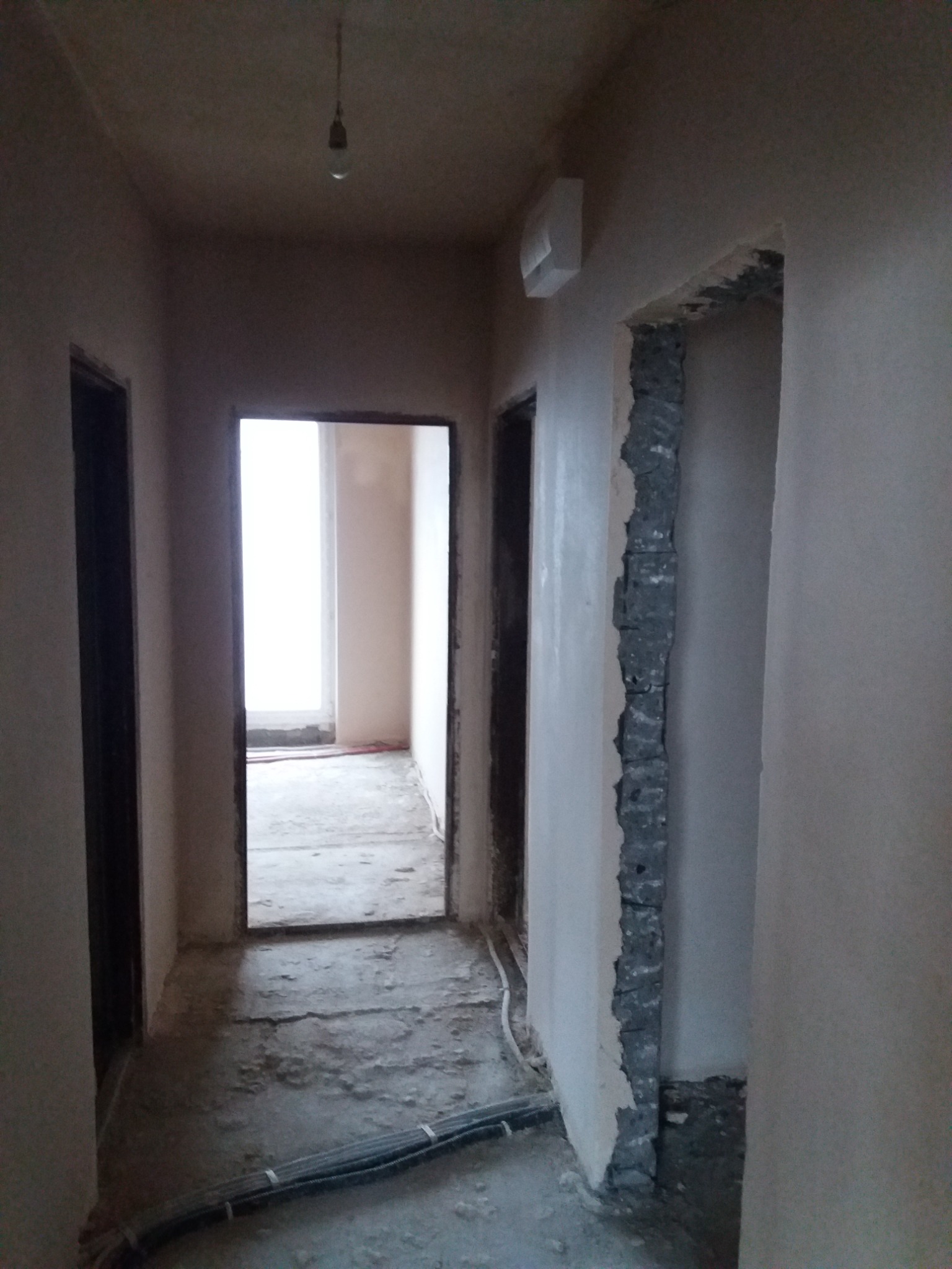 Korytarz w trakcie prac tynkarskich maszynowych; ściany pokryte świeżym tynkiem, widoczne kable na podłodze i surowa ościeżnica drzwiowa.