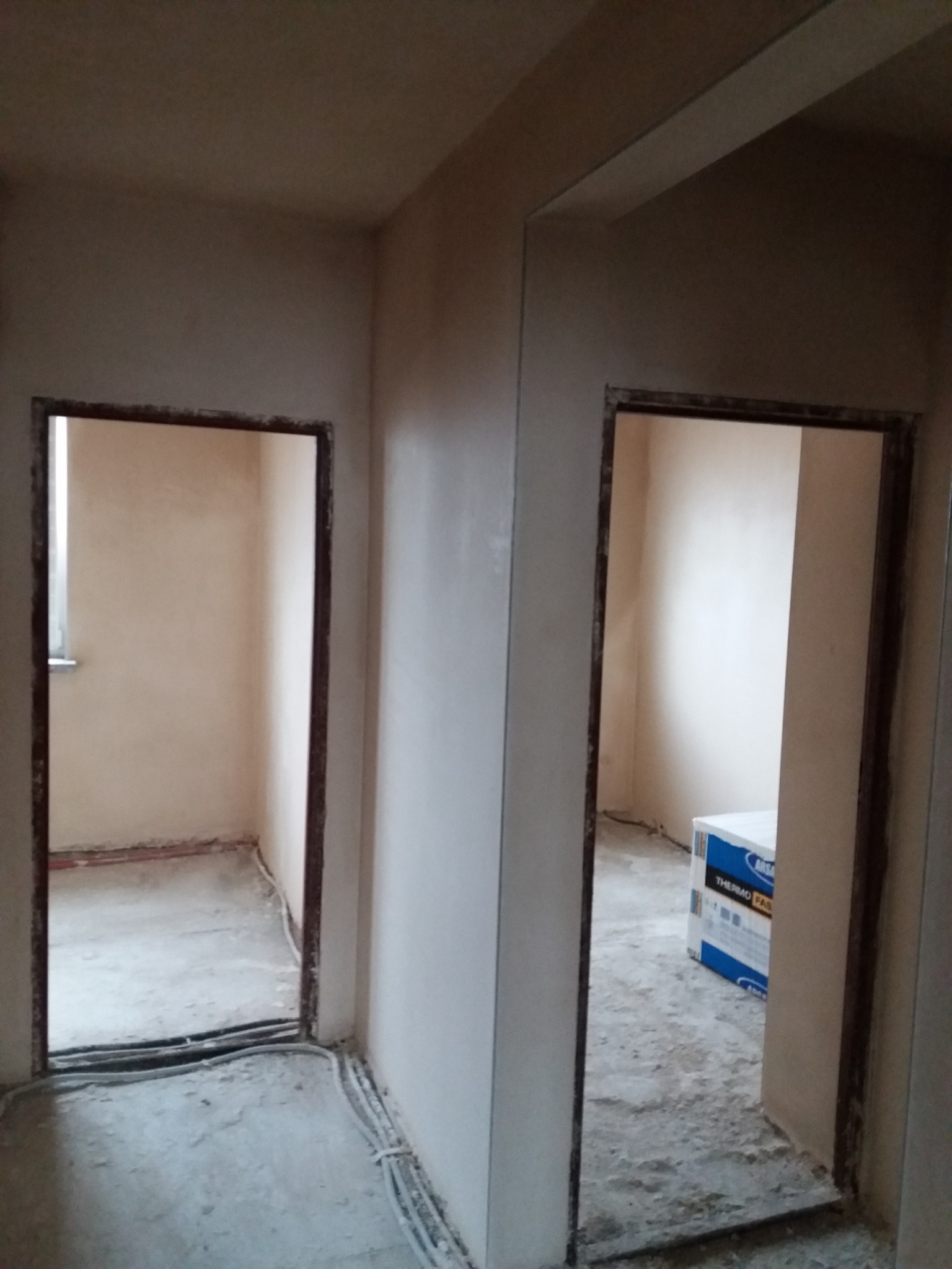 Wnętrze po nałożeniu gładzi, widoczne dwa otwory drzwiowe, przy jednym z nich stoi karton z materiałami budowlanymi.