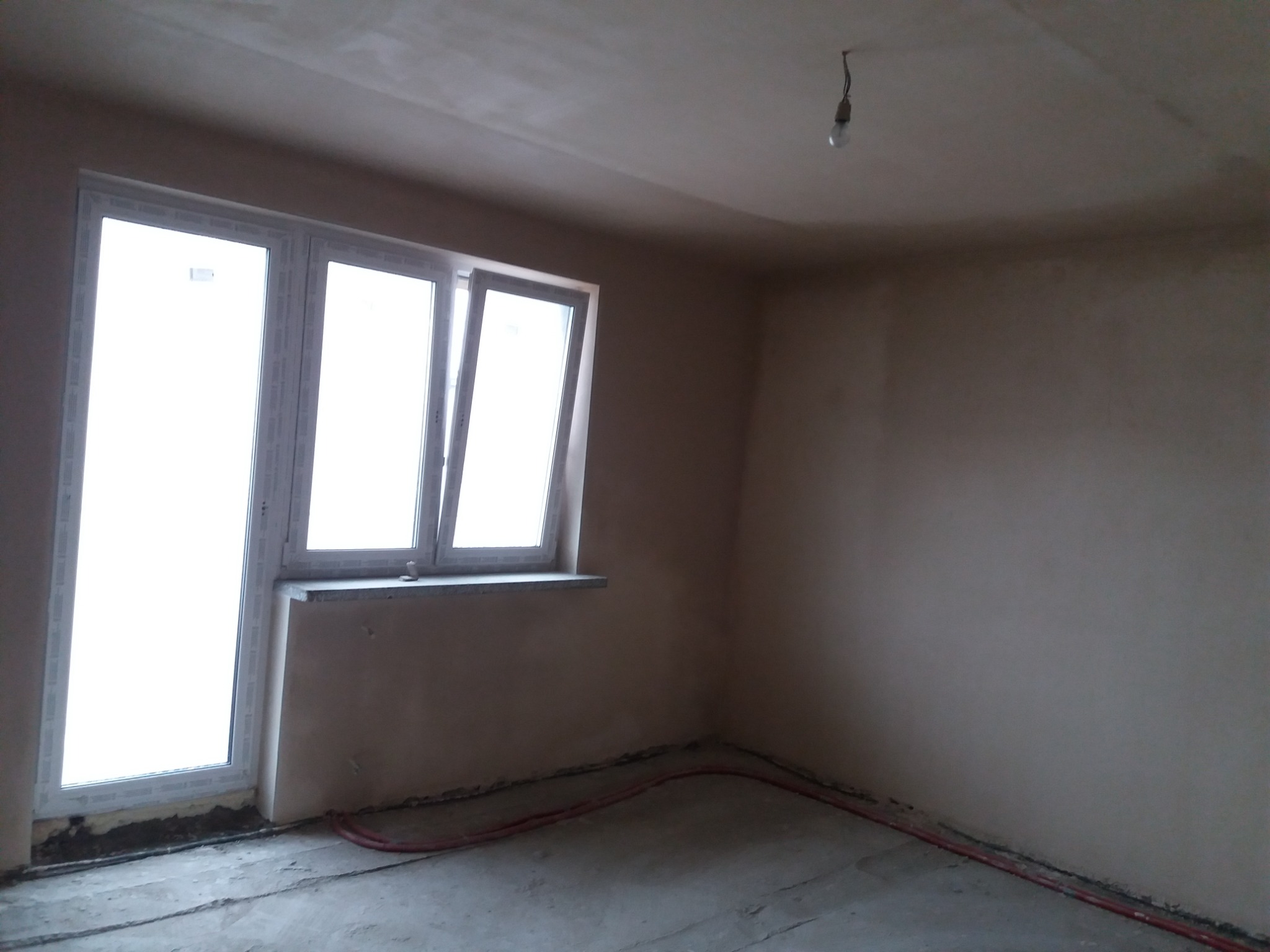 Pokój po nałożeniu tynków: ściany o jednolitej, jasnej powierzchni, widoczne okno z białymi ramami, podłoga w trakcie przygotowania, czerwony przewód na podłodze, surowa żarówka zwisająca z sufitu.