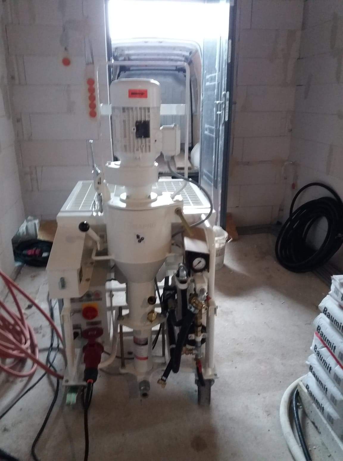 Maszyna do tynków maszynowych marki Dynapro, widoczna w surowym wnętrzu, z otwartymi drzwiami na zewnątrz, obok ułożone worki z zaprawą Baumit