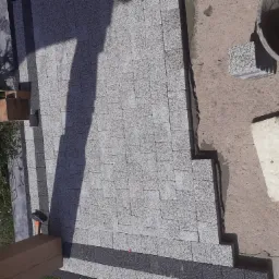 Układanie szarej kostki brukowej na podjeździe z widocznym fragmentem betonu i narzędziami murarskimi, częściowo zacienione