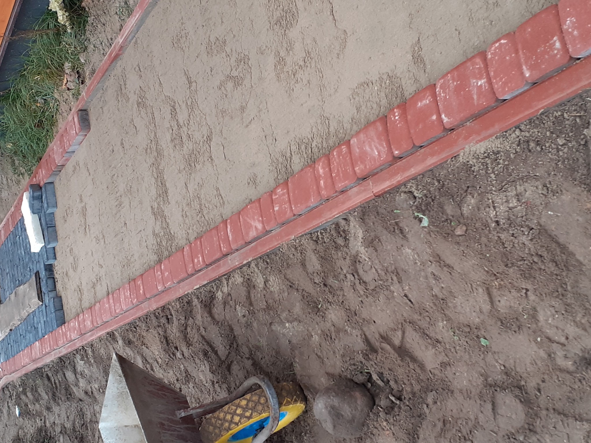 Układanie czerwonej kostki brukowej obramowującej teren, widoczne narzędzia i przygotowane podłoże z piasku.