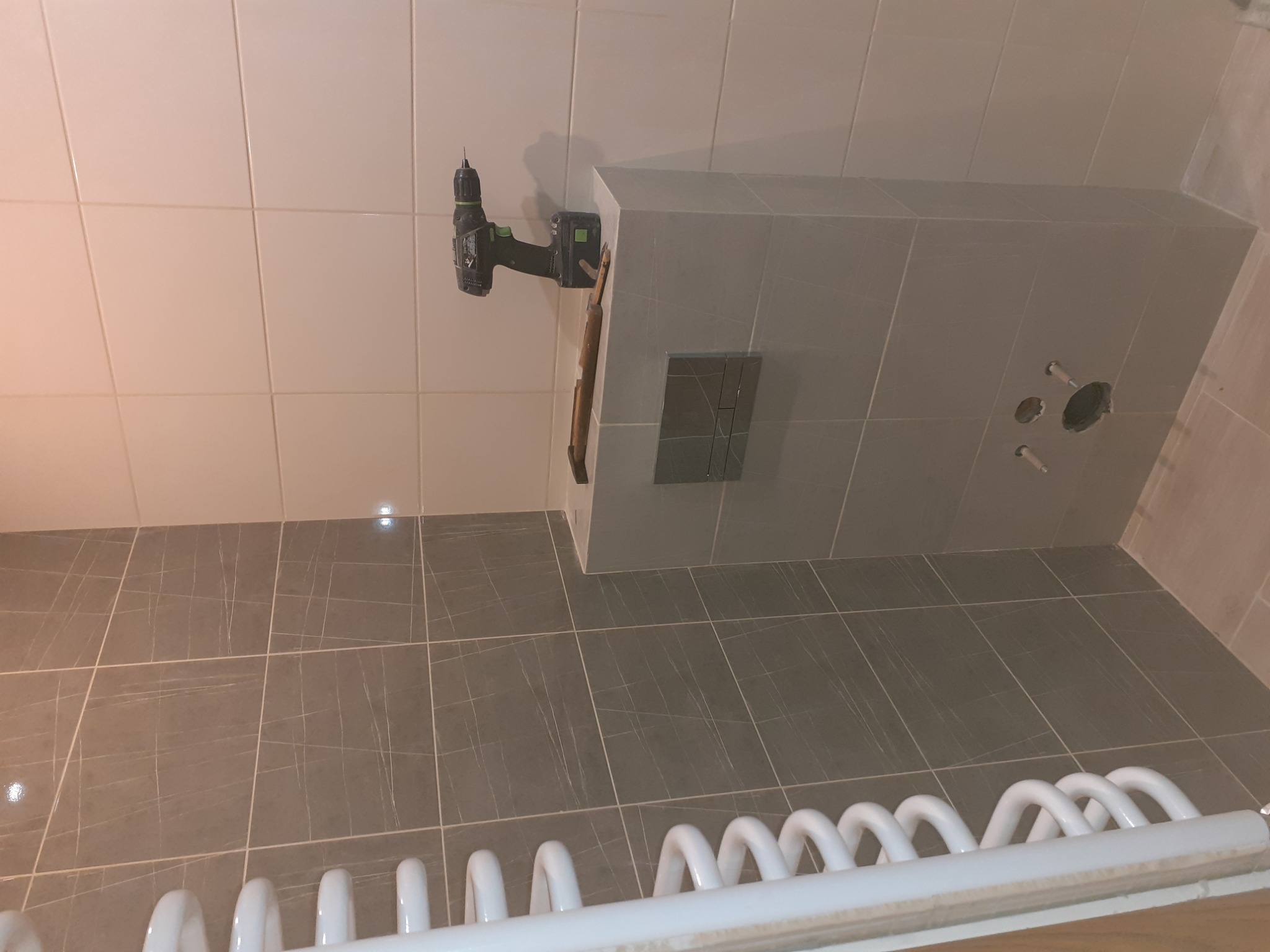 Szara łazienka w trakcie wykończenia z widocznym grzejnikiem drabinkowym, płytkami podłogowymi imitującymi marmur, oraz wiertarką akumulatorową zawieszoną na ścianie nad zabudową stelaża WC.