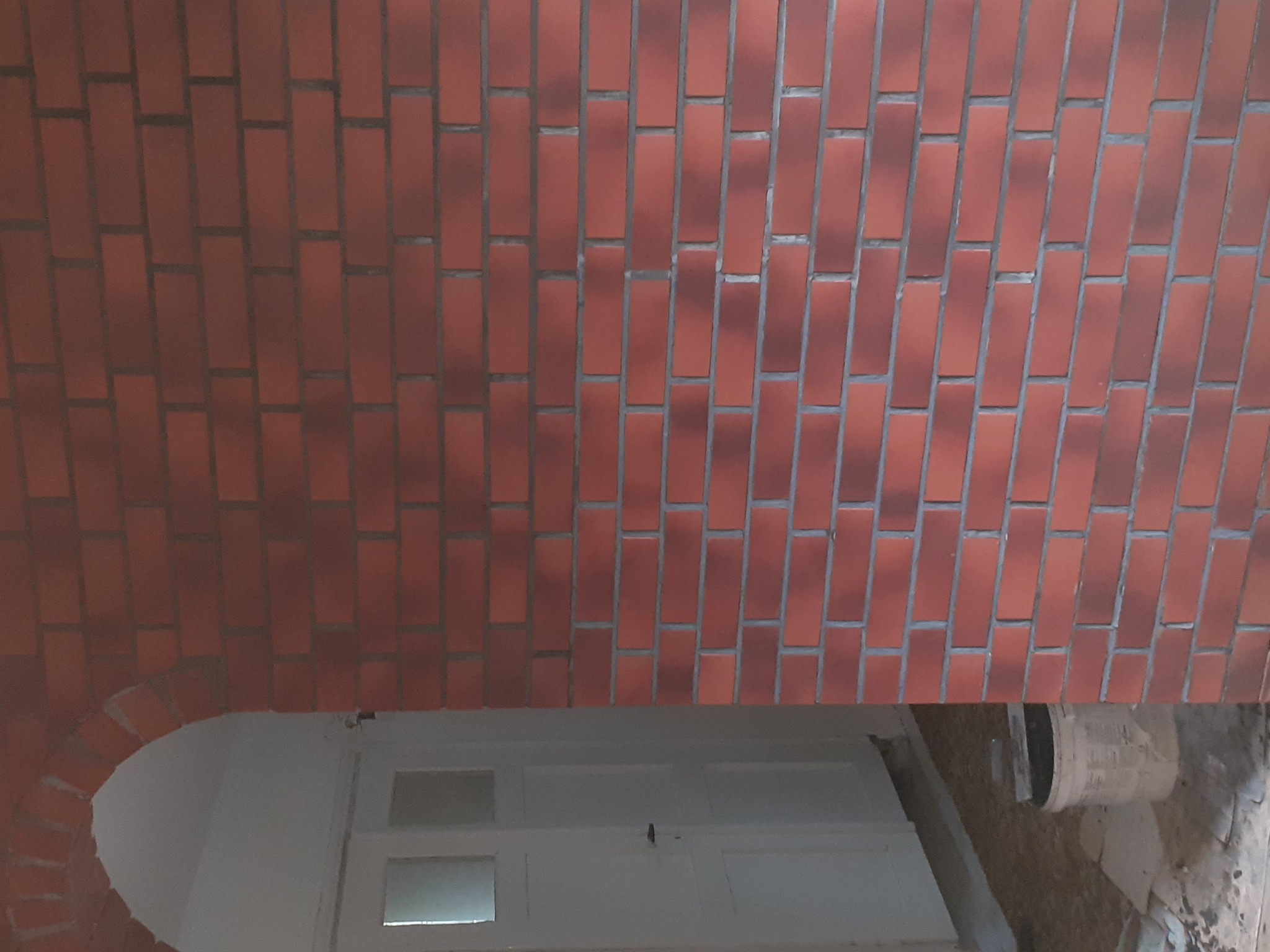 Ceglana ściana z łukowym przejściem, widoczny fragment białych drzwi i wiadro z zaprawą, ukośny kadr.