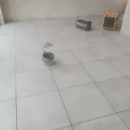 Wyłożona jasnymi, kwadratowymi płytkami podłoga w trakcie prac wykończeniowych; widoczne narzędzia: wiadro z zaprawą, paca do fugowania i grzejnik elektryczny.