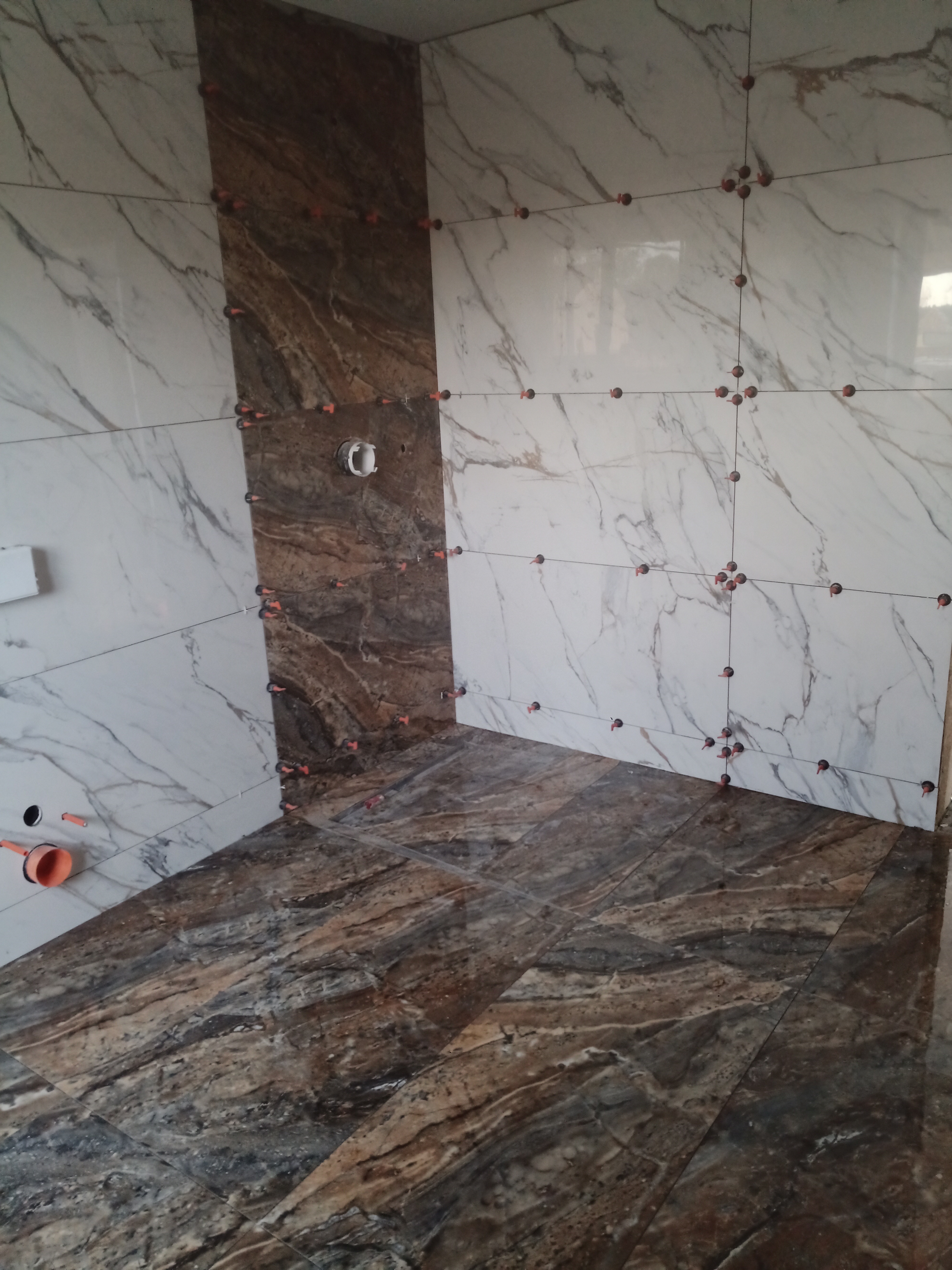 Wnętrze łazienki w trakcie układania płytek imitujących marmur w odcieniach bieli i brązu, widoczne krzyżyki dystansowe i otwór na instalację hydrauliczną.