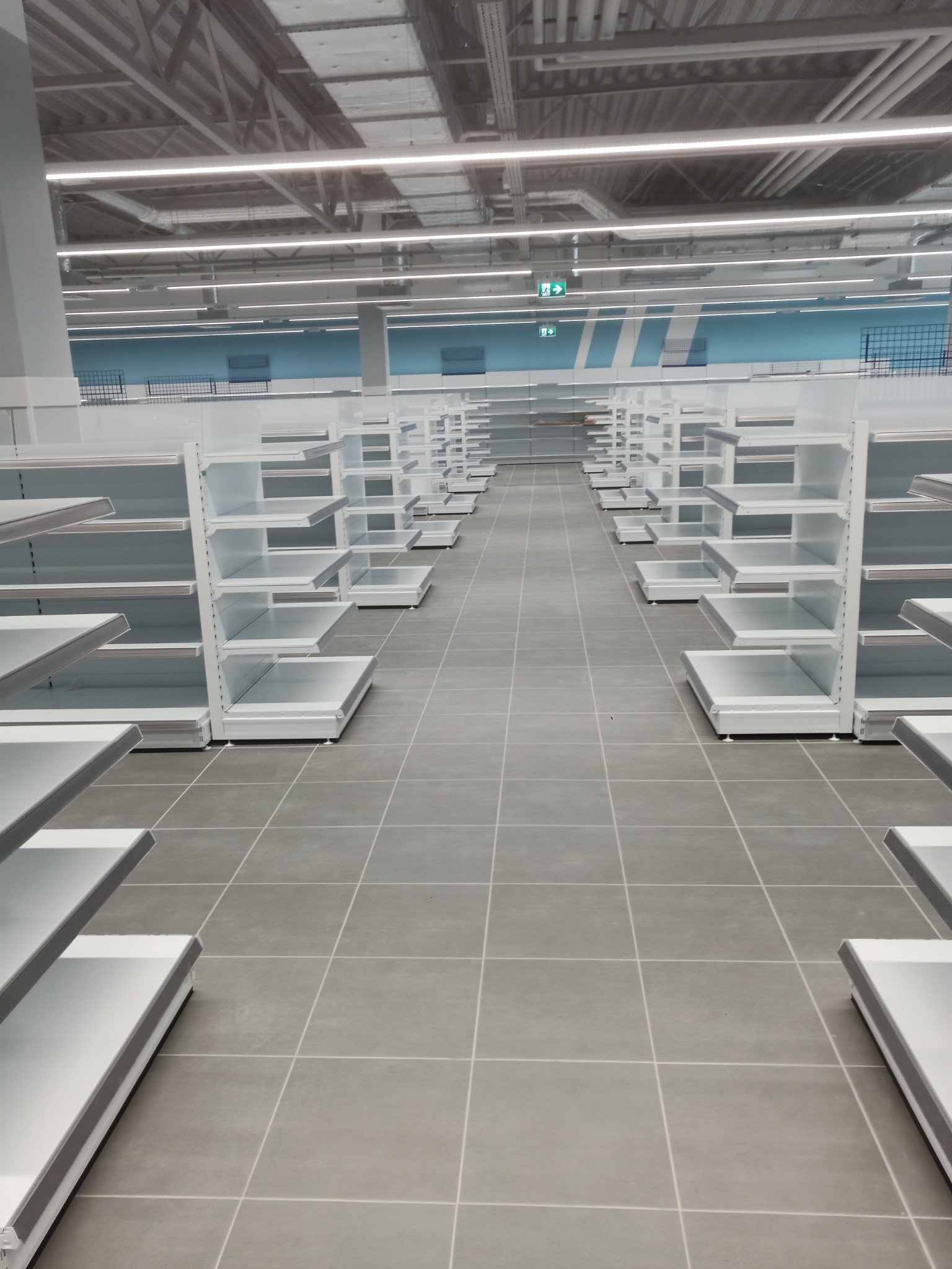 Długa perspektywa rzędu pustych, białych regałów sklepowych na szarych płytkach podłogowych, oświetlonych przez równomierne światło sufitowe, w tle błękitne akcenty na ścianach.