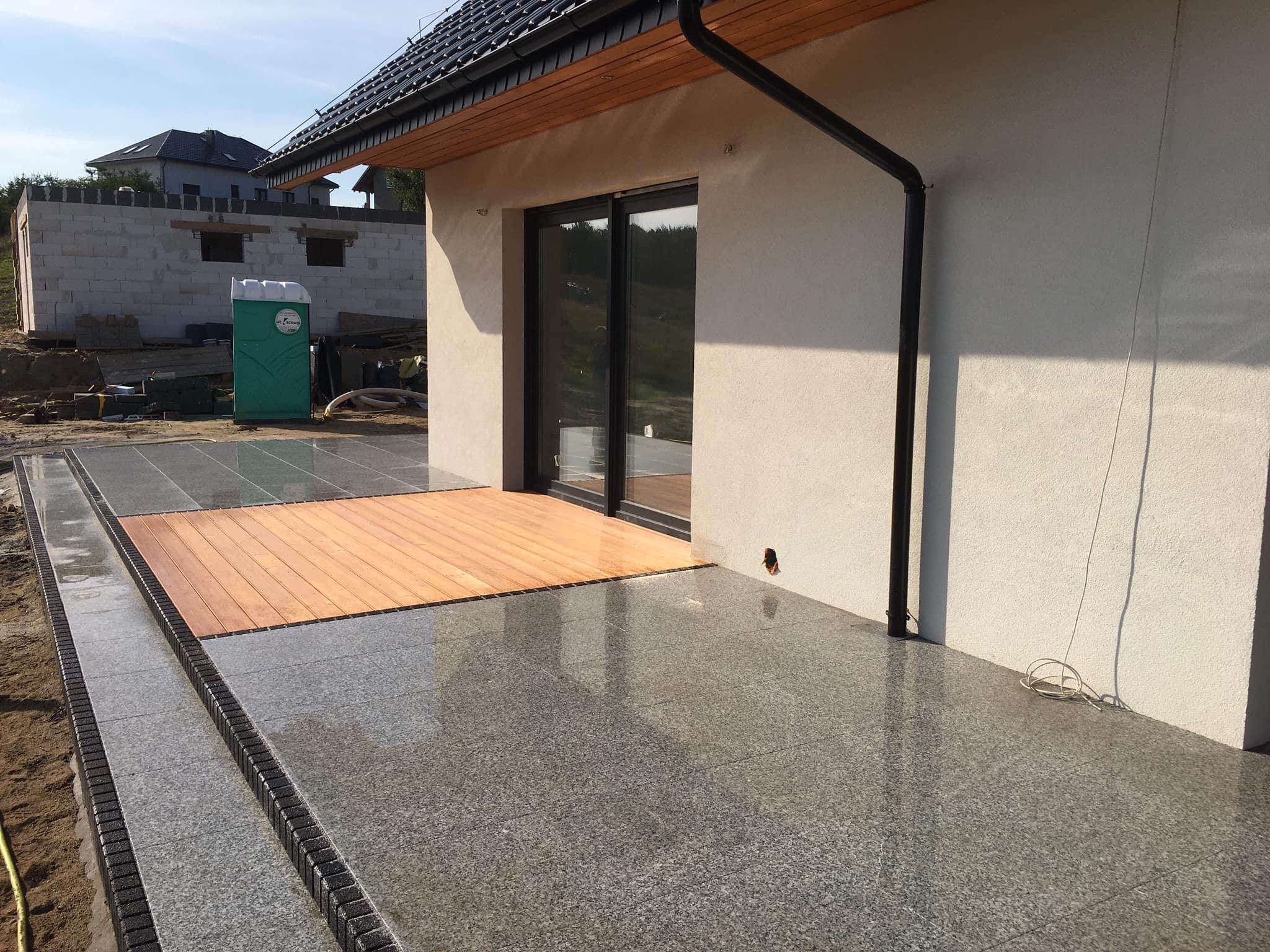 Granitowy taras z drewnianym podestem przylegający do budynku, widoczny fragment elewacji z czarną rynną i przesuwne drzwi tarasowe, w tle budynek w trakcie budowy i przenośna toaleta.