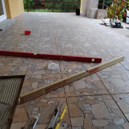 Układanie płytek na balkonie z użyciem poziomicy Sola+, nożyków do cięcia i krzyżyków dystansowych; widoczne zapasy płytek w tle.