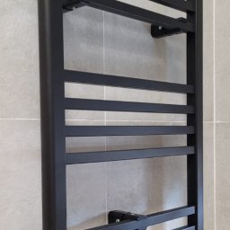 Plumber Crafting - Czarny, drabinkowy grzejnik łazienkowy zamontowany na szarej, kafelkowanej ścianie. Widoczne zawory termostatyczne i przyłącza. Minimalistyczny design.