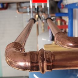 Plumber Crafting - Miedziane rury instalacji hydraulicznej z precyzyjnymi złączami, zamontowane pod sufitem, z widocznym zbiornikiem wyrównawczym i elementami mocującymi.