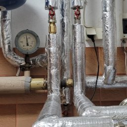 Plumber Crafting - Izolowane rury grzewcze z manometrem i zaworami, instalacja CO w kotłowni. Rury owinięte folią aluminiową, widoczne elementy instalacji i podłączenia.