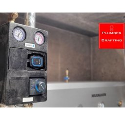 Czarna skrzynka z manometrami i pompą obiegową, element instalacji grzewczej na tle kotła. Widoczne rury i przyłącza. Szare tło ściany.