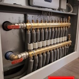 Rozdzielacz ogrzewania podłogowego w szafce, z widocznymi rurami, zaworami i sterownikiem elektronicznym. Widoczny logotyp firmy w prawym dolnym rogu.