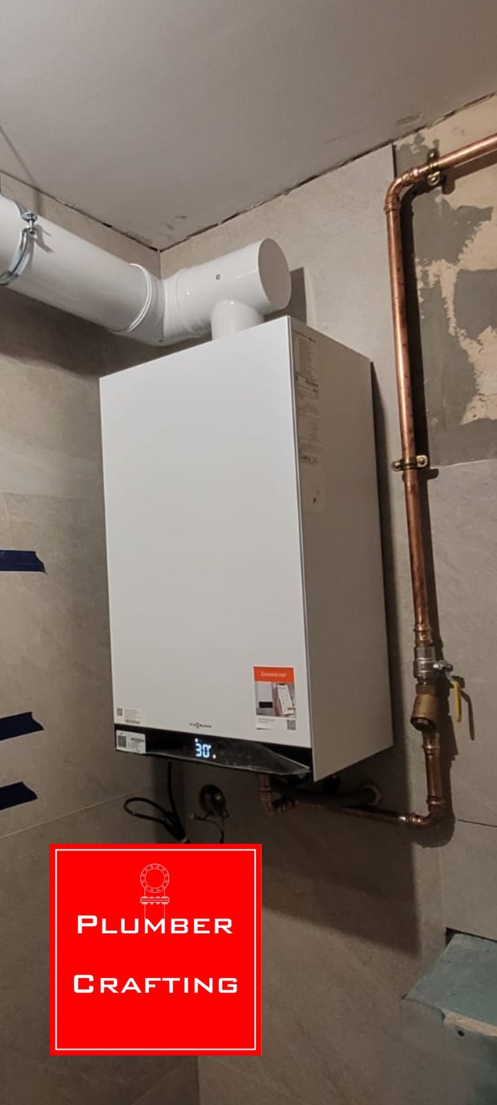 Biały piec gazowy Viessmann zamontowany na szarej, częściowo wykończonej ścianie z widocznymi rurami miedzianymi i białym przewodem wentylacyjnym.