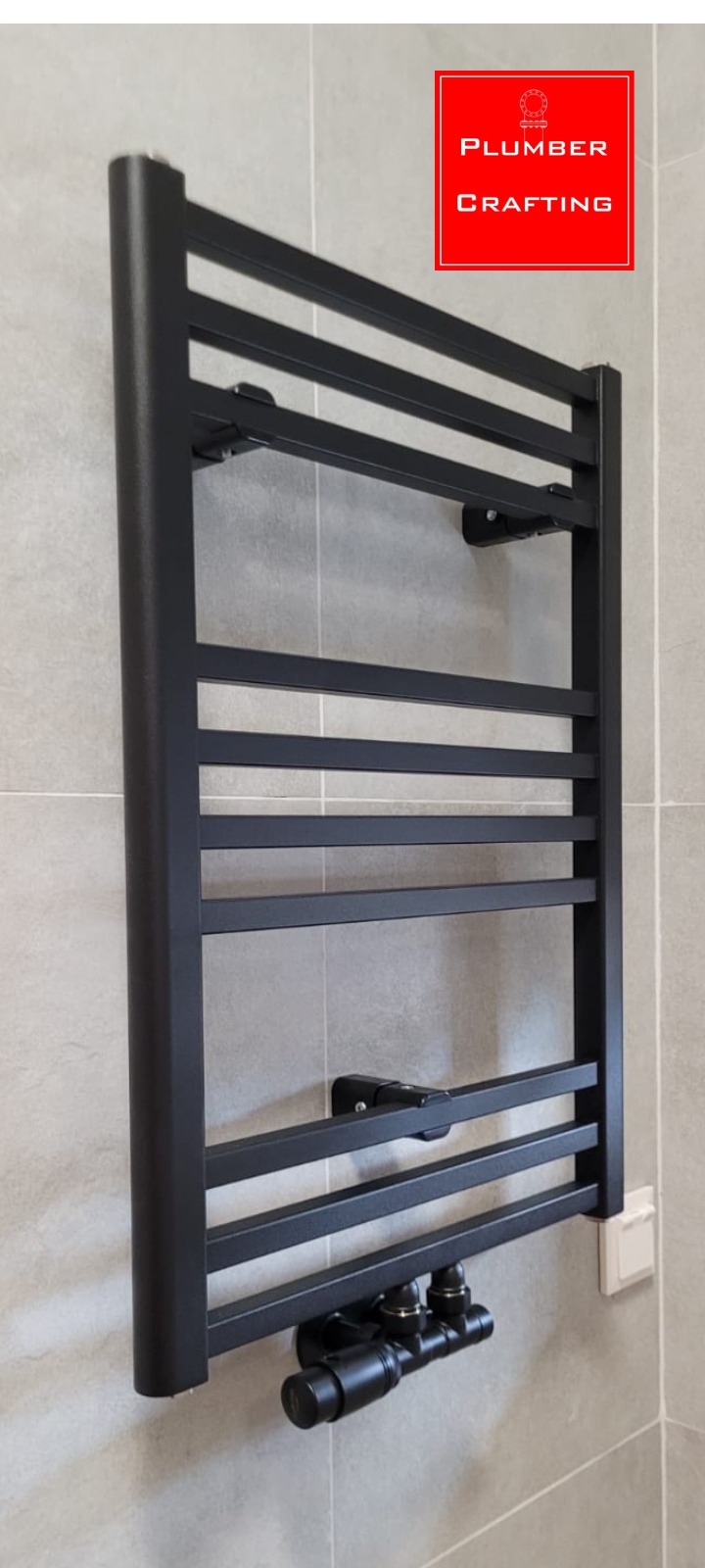 Czarny, drabinkowy grzejnik łazienkowy zamontowany na szarej, kafelkowanej ścianie. Widoczne zawory termostatyczne i przyłącza. Minimalistyczny design.