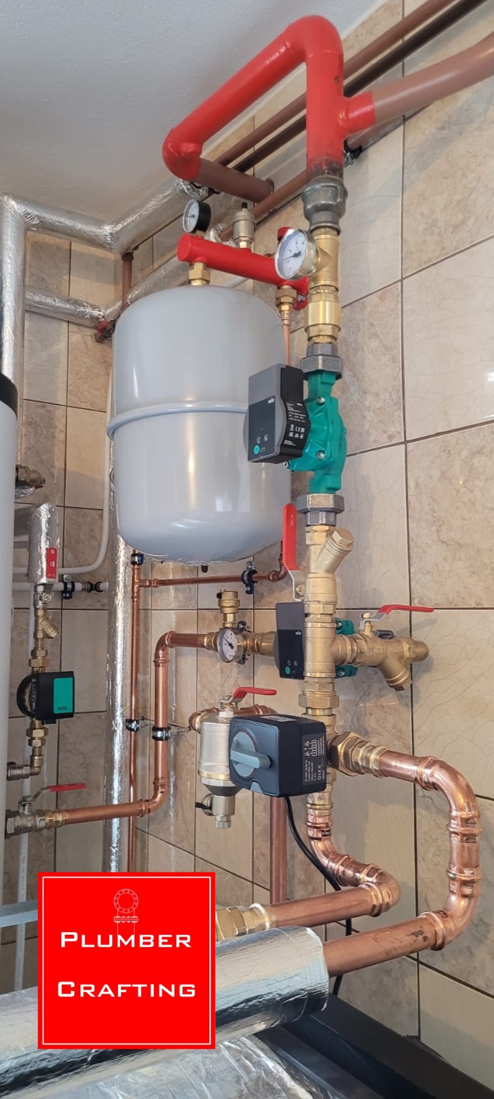 Złożona instalacja grzewcza z miedzianymi rurami, zaworami i manometrami na tle beżowych płytek. Widoczne elementy kotłowni i zbiornik wyrównawczy.