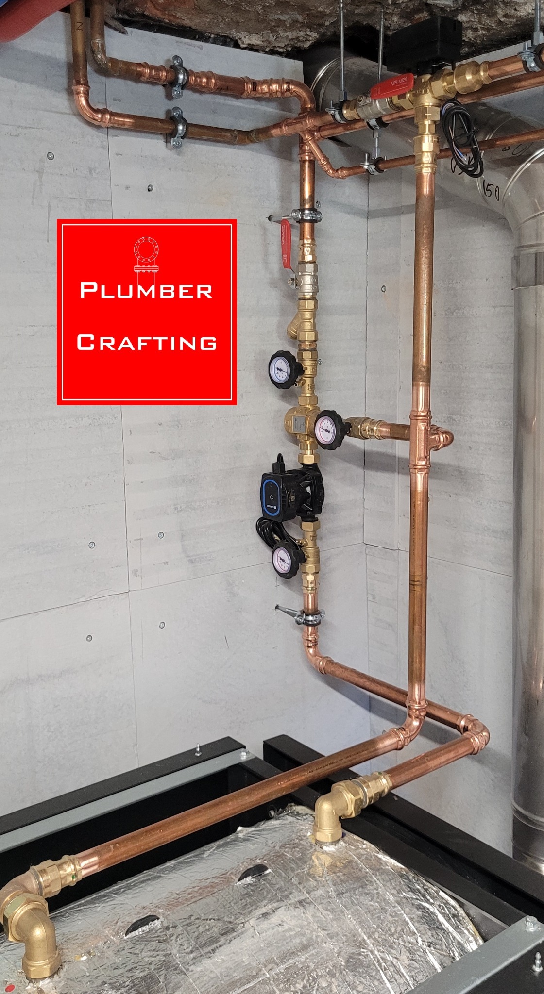 Miedziana instalacja grzewcza z manometrami i pompą obiegową, zamontowana na szarej ścianie. Widoczne rury, zawory i elementy mocujące. Część instalacji jest zaizolowana.