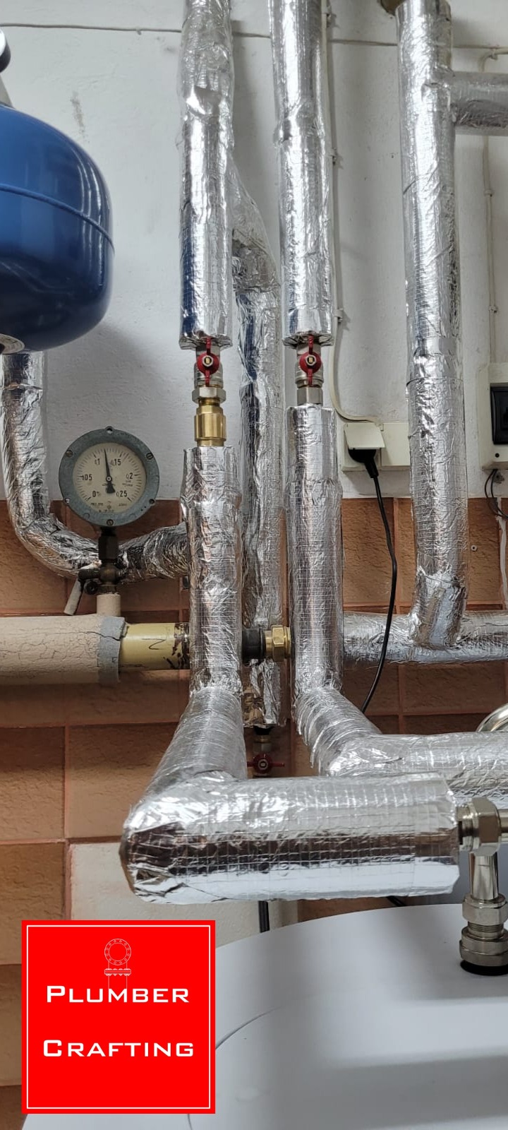 Izolowane rury grzewcze z manometrem i zaworami, instalacja CO w kotłowni. Rury owinięte folią aluminiową, widoczne elementy instalacji i podłączenia.