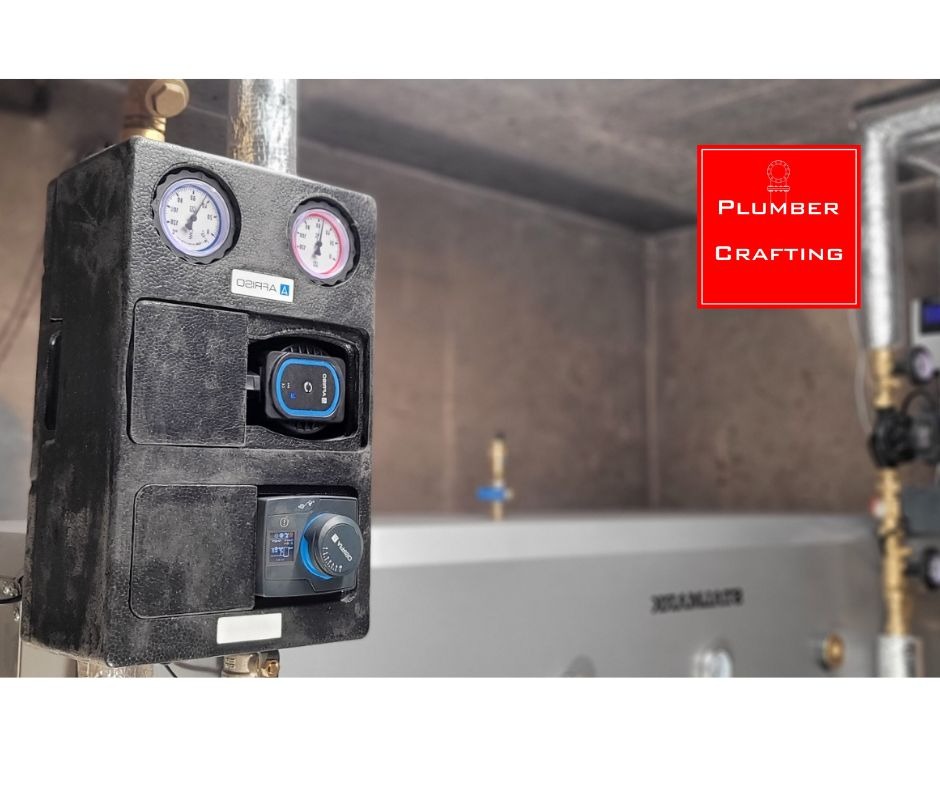 Czarna skrzynka z manometrami i pompą obiegową, element instalacji grzewczej na tle kotła. Widoczne rury i przyłącza. Szare tło ściany.