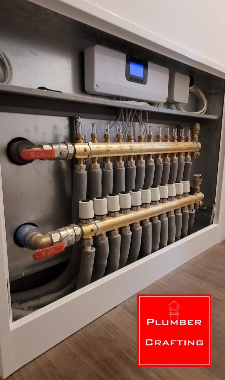 Rozdzielacz ogrzewania podłogowego w szafce, z widocznymi rurami, zaworami i sterownikiem elektronicznym. Widoczny logotyp firmy w prawym dolnym rogu.