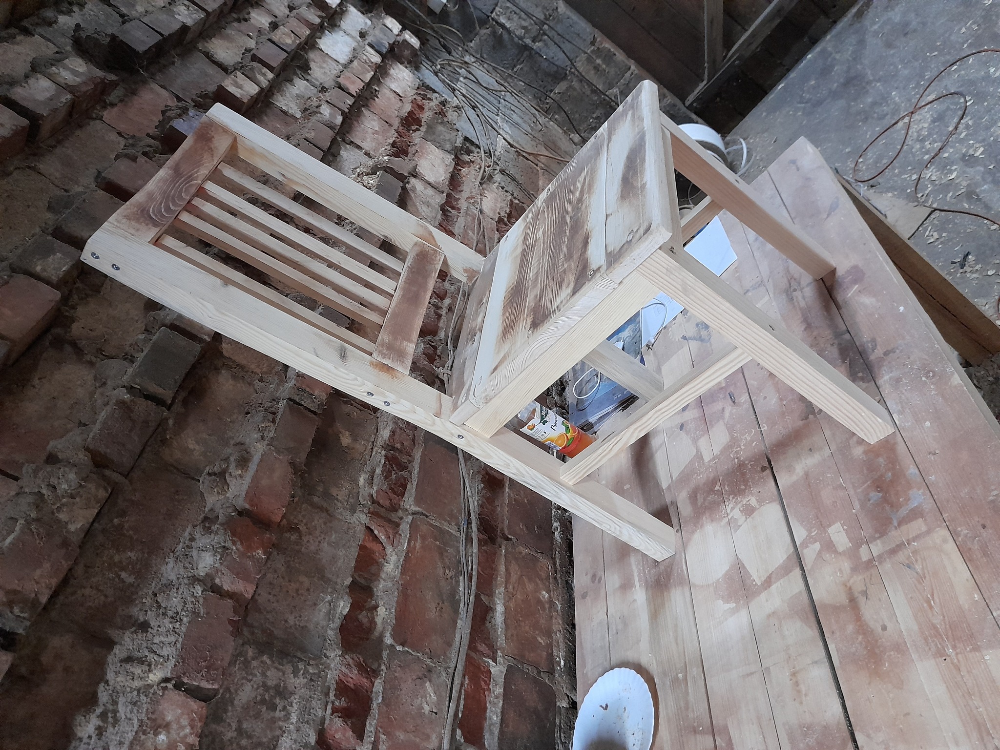 Surowe drewniane krzesło z oparciem szczebelkowym, częściowo wykończone, leżące na ceglanej podłodze w trakcie prac stolarskich, widoczne przewody i resztki materiałów budowlanych.