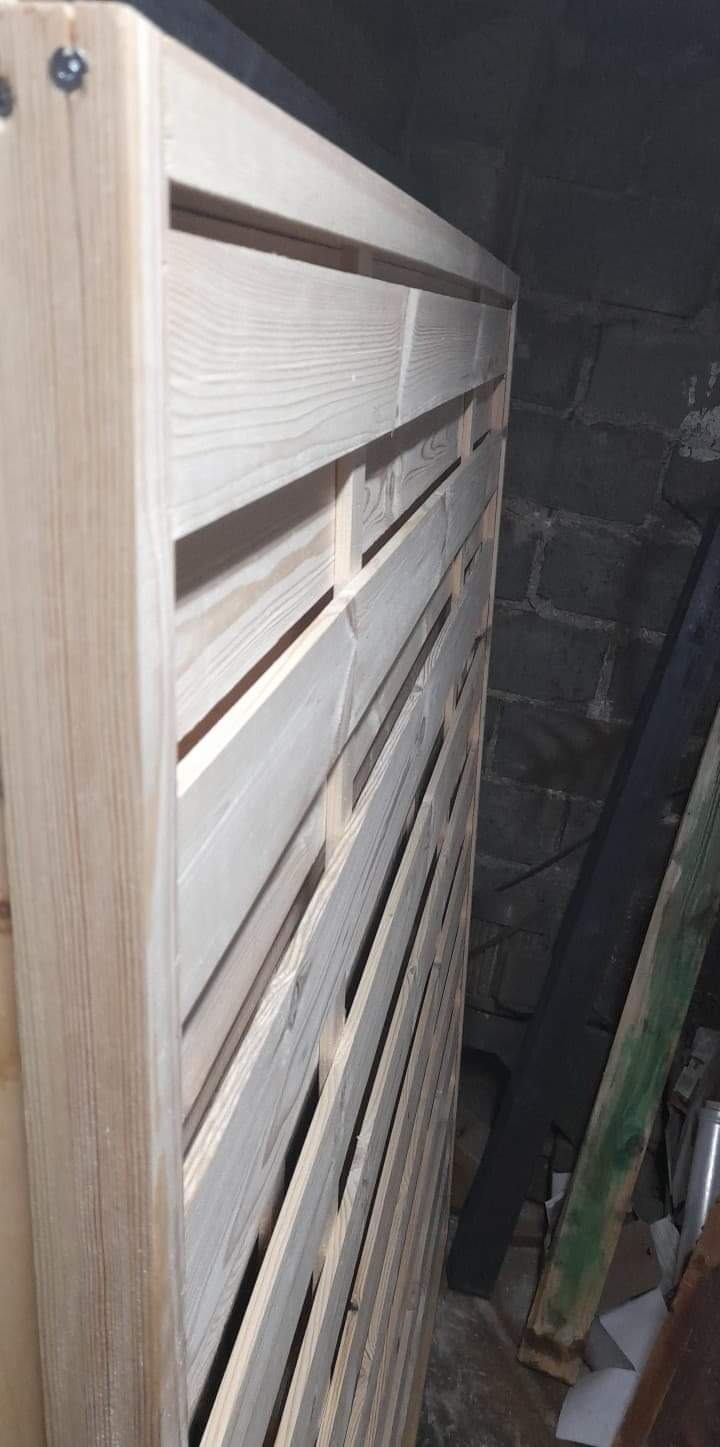 Surowe drewniane ogrodzenie panelowe z widocznymi szczelinami, montowane przy ciemnej, ceglanej ścianie, w surowym otoczeniu budowlanym.