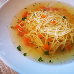 Rosół z makaronem nitki, marchewką i zieloną pietruszką w białym talerzu, ustawionym na drewnianym blacie stołu.