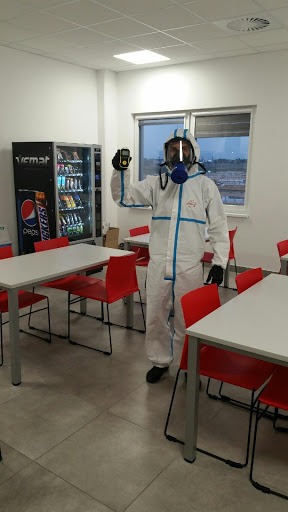 Pracownik w kombinezonie ochronnym i masce, trzymający miernik w dłoni, stoi w pomieszczeniu w Poznaniu. W tle widoczny automat z napojami i przekąskami oraz stoliki z czerwonymi krzesłami.