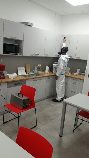 Pracownik w kombinezonie ochronnym i masce mierzy poziom czegoś przy szafkach kuchennych, obok stoi urządzenie na czerwonym krześle.