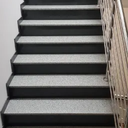 Schody wewnętrzne z szarego kamienia z ciemnymi krawędziami, z metalową balustradą i rośliną doniczkową na górnym stopniu.