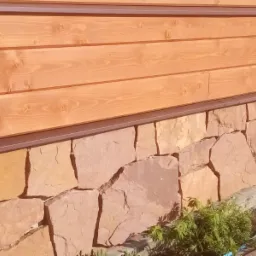 Wykończenie elewacji domu: drewniana boazeria w ciepłym odcieniu brązu zestawiona z rustykalnym, kamiennym cokołem z nieregularnych, piaskowych płyt. Widoczne zielone rośliny przy podstawie cokołu.