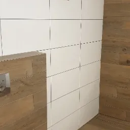 Wykończona łazienka w Inowrocławiu z białymi i drewnopodobnymi płytkami, widoczny fragment okna z profilem Salamander oraz zabudowa stelaża WC z drewnopodobnych płytek.