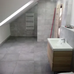 Wykończona łazienka na poddaszu z szarymi płytkami, drewnianą szafką pod umywalką i grzejnikiem drabinkowym, widoczna poziomica oparta o ścianę.