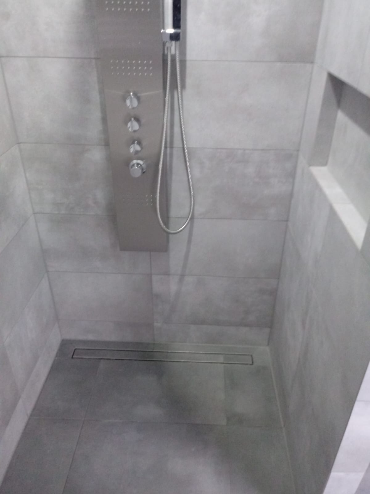 Szara kabina prysznicowa z panelem natryskowym i odpływem liniowym, wykończona płytkami w odcieniach szarości, z wnęką w ścianie.